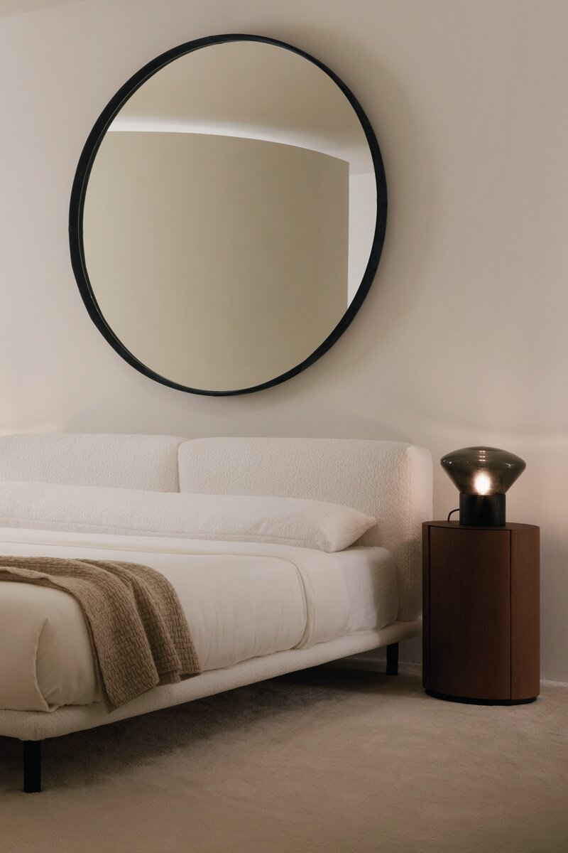 Round mirror on a wall above a bed with a side table and lamp.