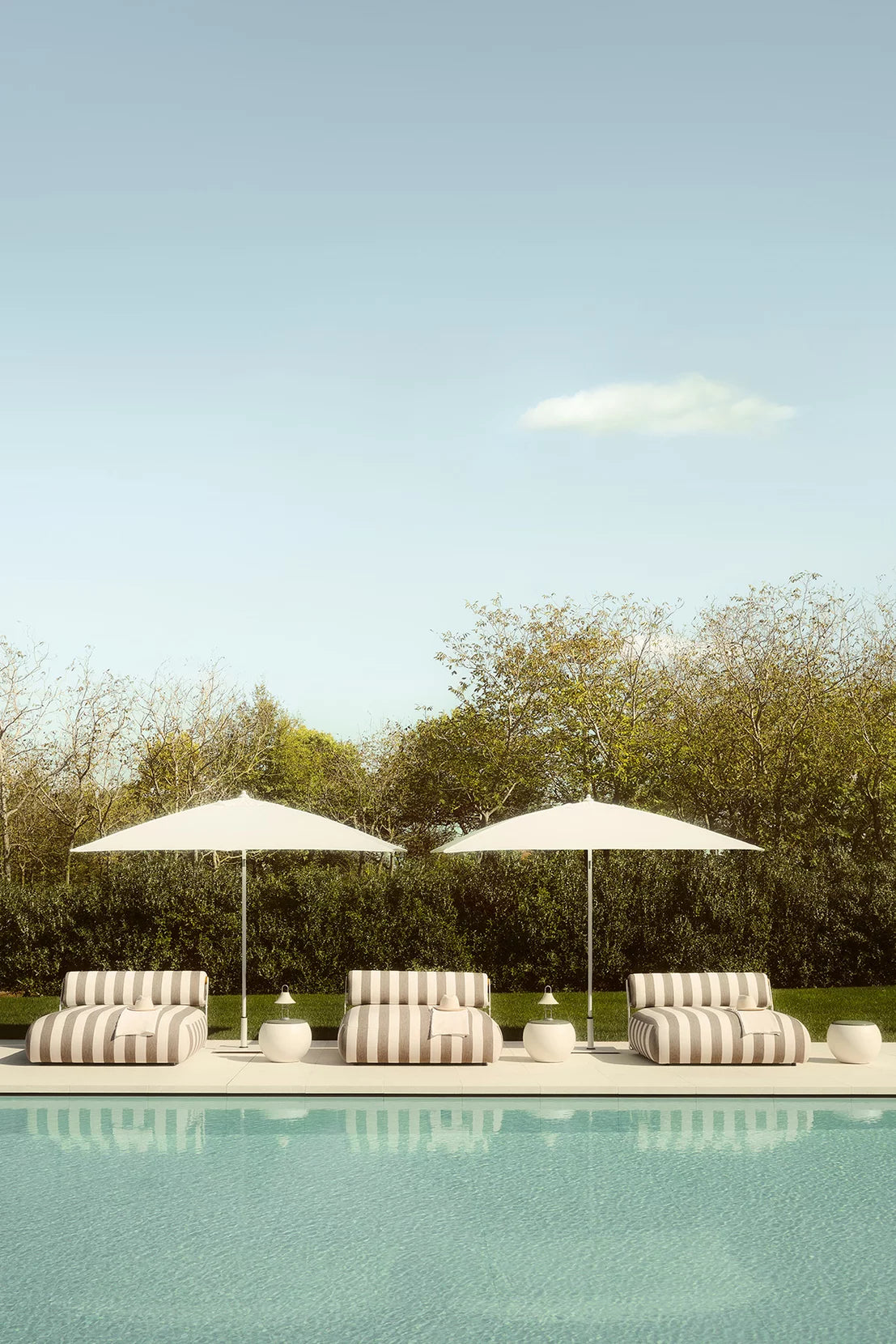 Three striped lounge chairs under white umbrellas by a pool with a clear sky.